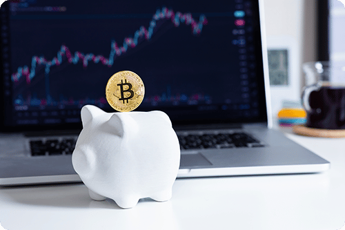 A small white piggy bank in front of a laptop showing a line graph with increasing returns. A golden bitcoin is entering the piggy bank.